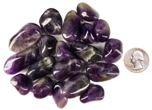 A pile of large amethyst tumbled stones with deep purple, to violet hues, and some white patterns, with a quarter for comparison. Some are smaller than the quarter, and some the samdiameter or larger.