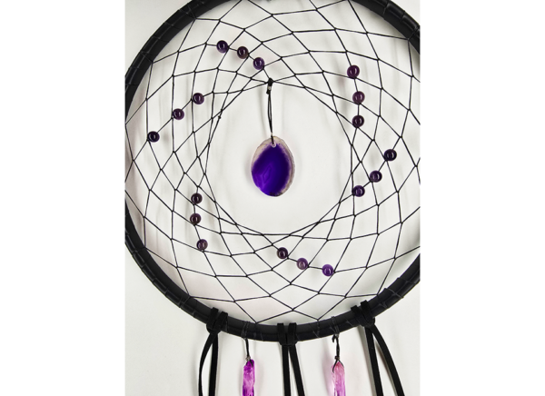 A dream catcher showing a purple agate slice as the centerpiece, surrounded by amethyst beads woven into the dream catcher, and purple quarts crystals hanging bellow.