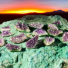 A group of amethyst geode clusters with deep purple crystals. They are displayed on a green rock with a sunset in the background.