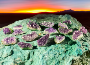 A group of amethyst geode clusters with deep purple crystals. They are displayed on a green rock with a sunset in the background.
