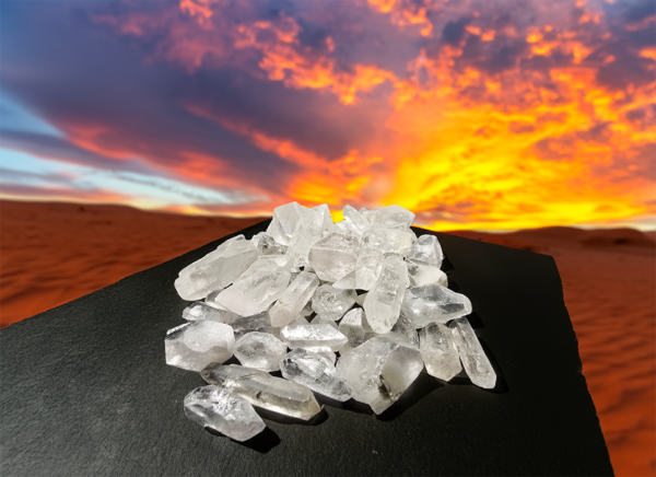 A pile of clear quartz crystals on display on a black table in the desert with a sunet in the background.