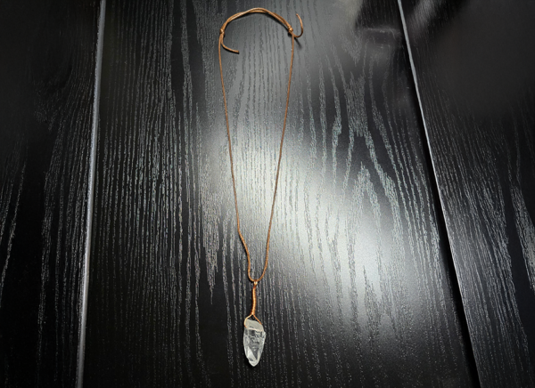A quartz pendant with a copper wire wrapped bail with a wax cord adjustable necklace displayed on a black table.