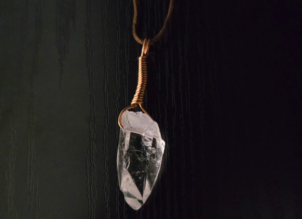 A quartz crystal pendant drilled that has a copper wire wrapped bail, with a black background showing the clarity of the crystal.