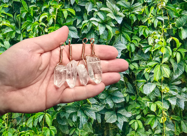 Four quarts pendants with wire wrapped copper bail in the palm of a hand outdoors in front of a bush.