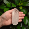 A large clear quartz point in the palm of a hand with a green, leafy background. It is quite large. Labeled 'J'.