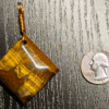 A yellow brown tiger's eye cabochon square shaped with a copper wire bail, and with a quarter for size comparison. It is a bit larger than the quarter.
